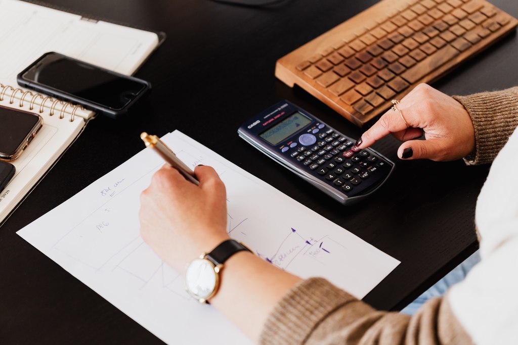 Pessoa escrevendo em folha de papel e usando calculadora, simbolizando imposto de renda 2023. Também um teclado de computador, um caderno e celulares sobre a mesa